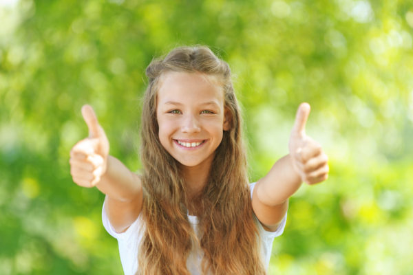 Beautiful teenage girl in white blouse lifts thumb upwards, against green of summer park.
